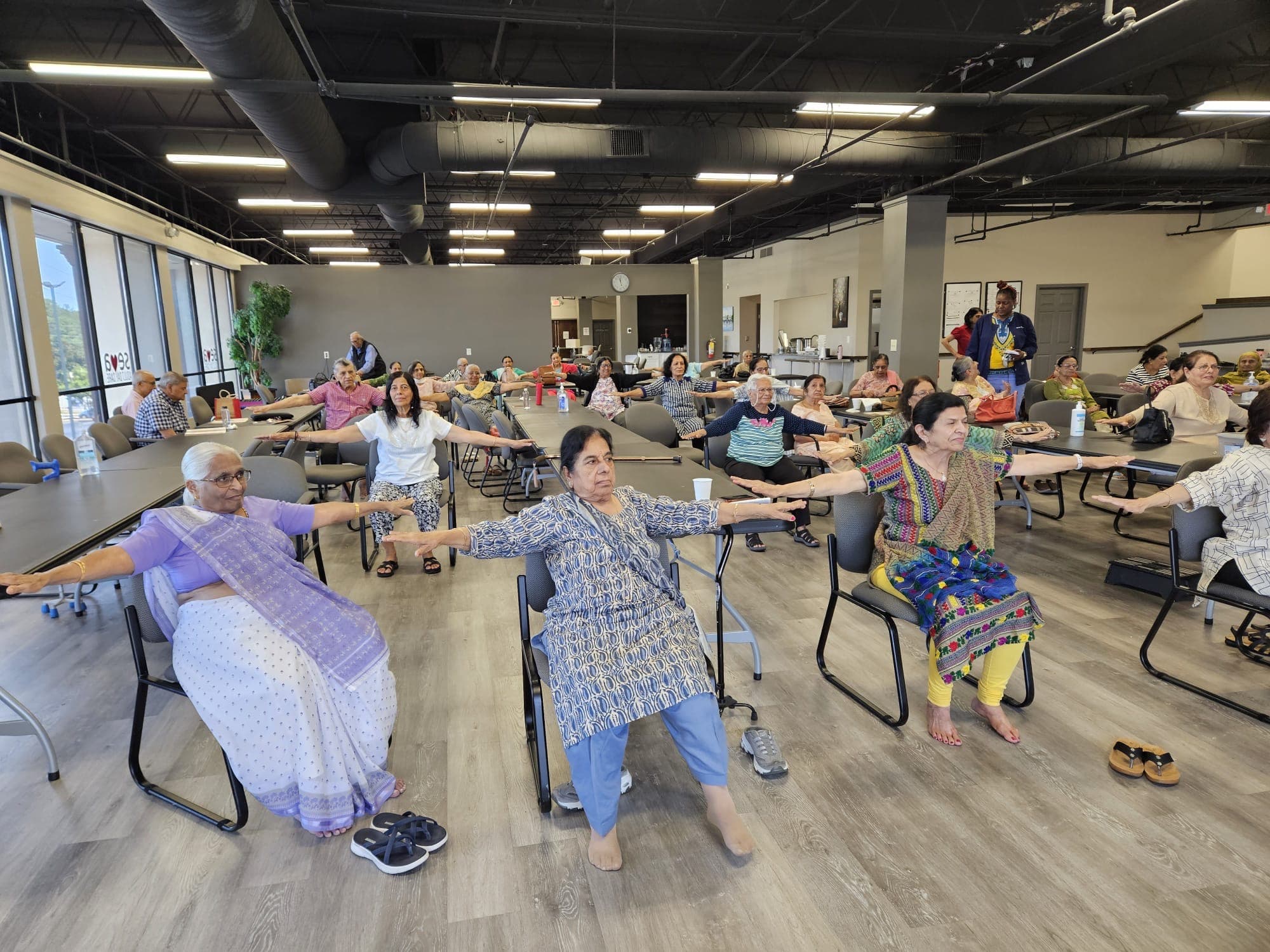 Members of the Seva Senior Day Center in a seated chair yoga class, arms outstretched